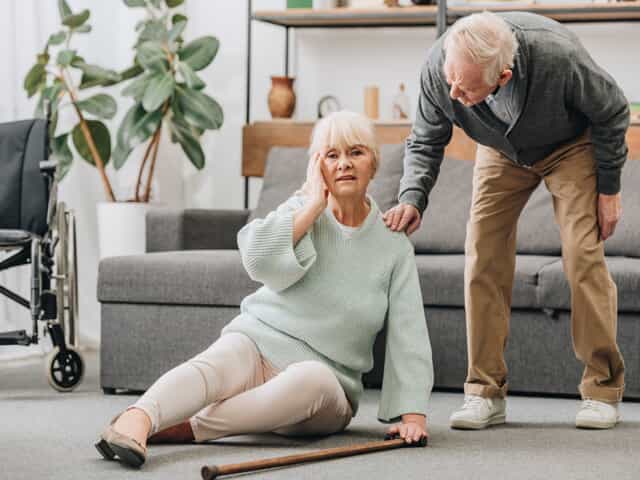 Senior lady on the ground with hand to her head after falling. Man bending over to check on her wellbeing.
